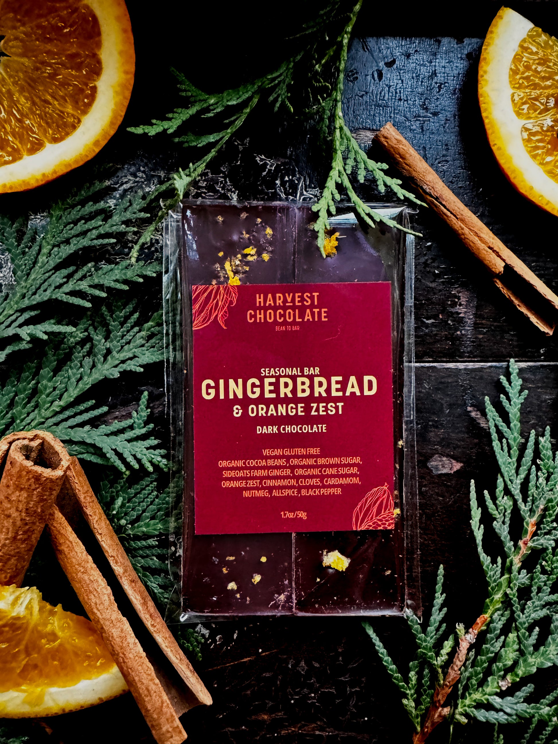 A Gingerbread + Orange Zest dark chocolate bar from Harvest Chocolate, surrounded by orange slices and pine branches on a dark wooden surface.