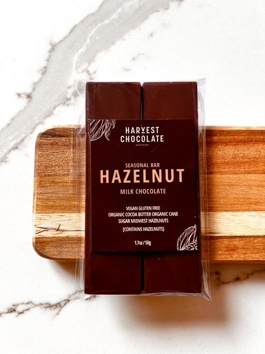 A Hazelnut Milk Chocolate bar, labeled "Harvest Chocolate," is wrapped in clear packaging and rests on a wooden surface against a marble background. The bar is vegan, gluten-free, and made