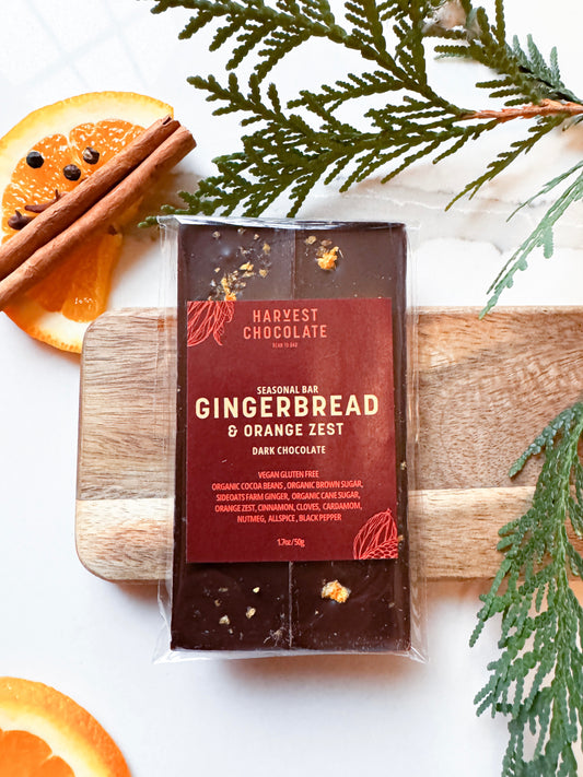 A bar of Harvest Chocolate Gingerbread + Orange Zest chocolate on a wooden board, surrounded by dried orange slices and pine branches. The packaging displays ingredients and the flavor profile.