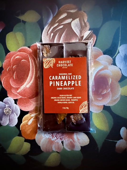 A bar of Harvest Chocolate's Caramelized Pineapple dark chocolate on a floral-patterned background. The package features clear branding and product ingredients, with visible slices of pineapple brittle.
