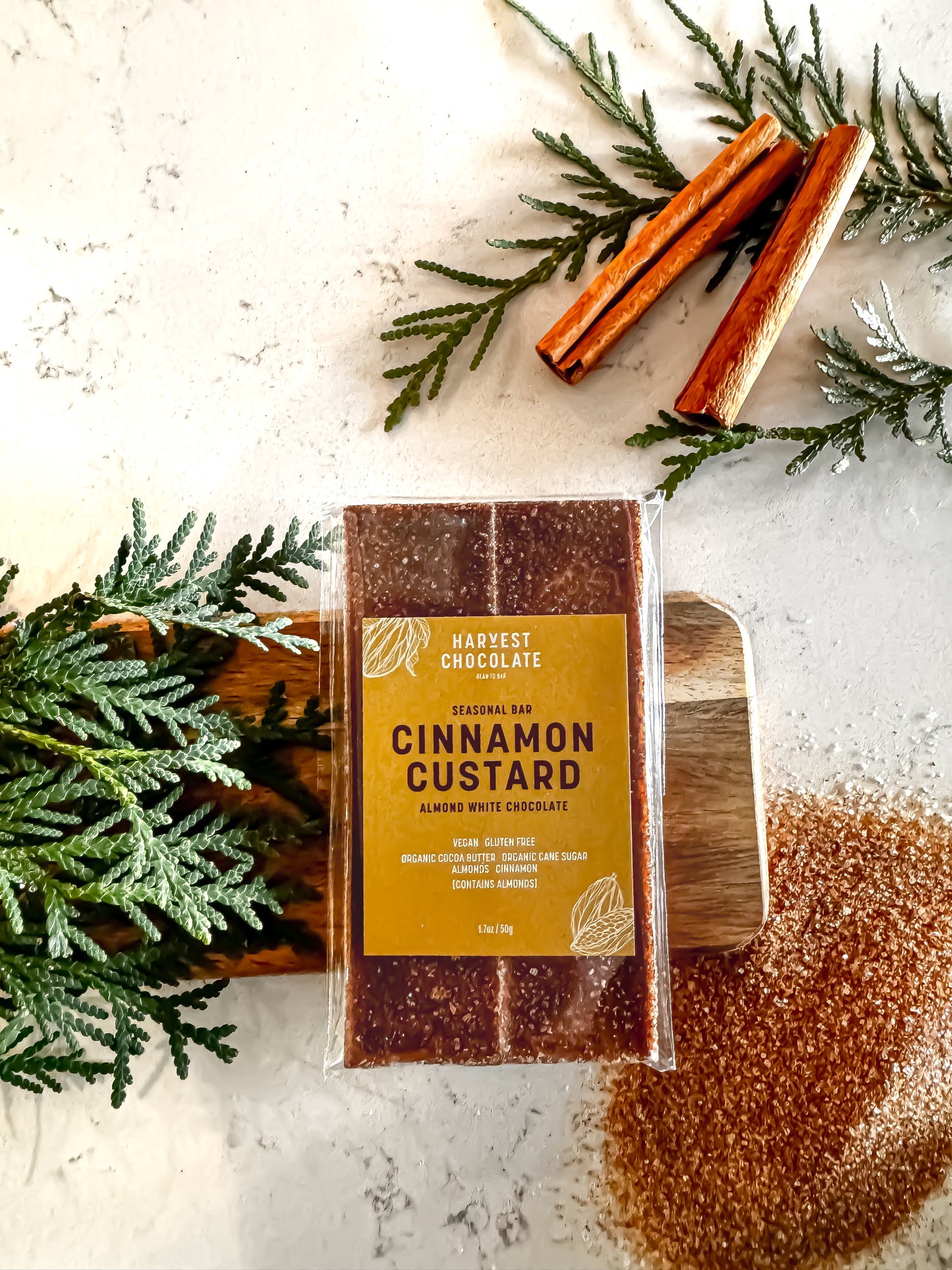 A bar of Harvest Chocolate's classic Cinnamon Custard chocolate placed on a wooden spatula, surrounded by scattered cinnamon sticks, sprinkled cocoa, and green fir branches on a light surface.