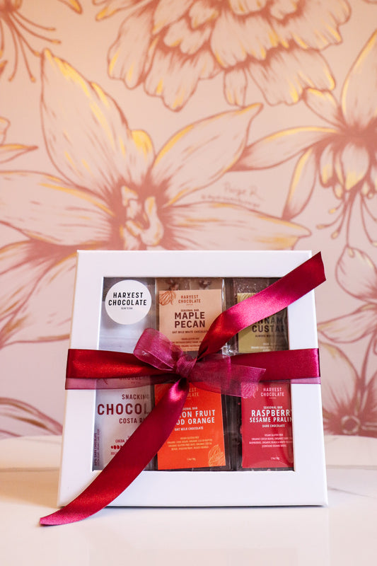 A Harvest Chocolate Gift Box wrapped with a red ribbon contains various craft chocolates, including flavors like maple pecan and raspberry sesame, against a floral wallpaper background.