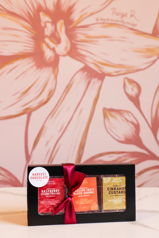 A Small Batch Chocolate Gift Box from Harvest Chocolate with raspberry, passion fruit, and cinnamon flavors, tied with a red ribbon, sits in front of a floral printed background.