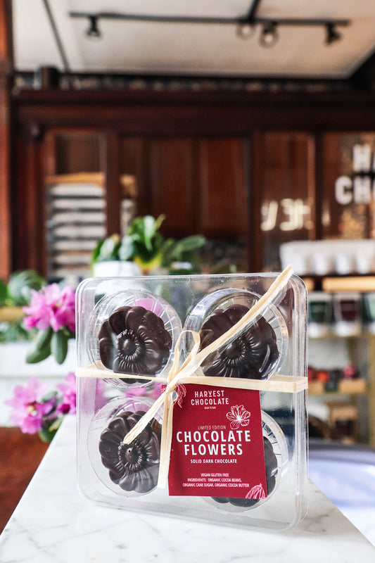A pack of Harvest Chocolate Flowers made from Ecuadorian cocoa beans on a marble countertop with "Harvest Chocolate Company" branding, tied with a gold ribbon, placed in front of a floral café background.