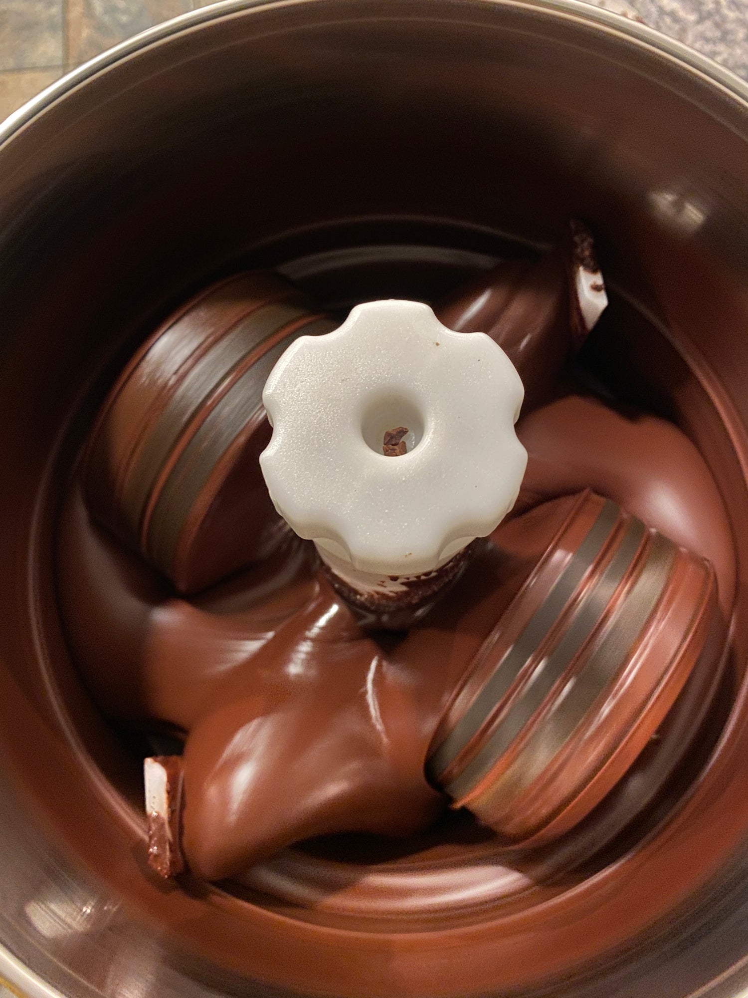 A white and chocolate swirl bundt cake sits in a rotating tin, partially coated with glossy, melting chocolate that is being evenly spread over its surface.
