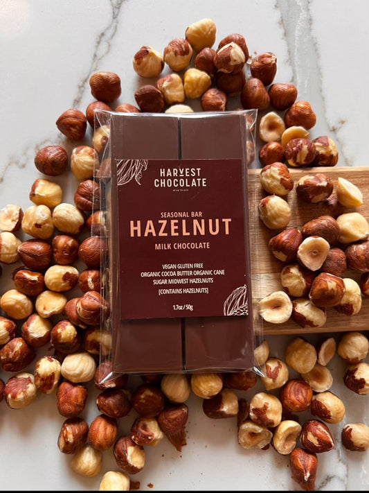 A Midwest grown Harvest Chocolate hazelnut milk chocolate bar in a brown wrapper is surrounded by scattered whole hazelnuts on a marble surface. The wrapper has white and gold text detailing the product's ingredients and organic.