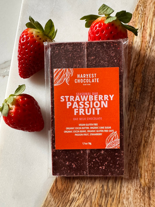 A bar of Harvest Chocolate milk chocolate with "Strawberry Passion Fruit" flavor, prominently displayed on a white marble surface, surrounded by fresh strawberries. The packaging is orange with decorative patterns.