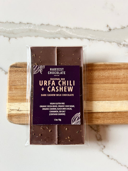 A packaged chocolate bar labeled "Urfa Chilli + Cashew Dark Cashew Milk Chocolate" from Harvest Chocolate, placed on a wooden roller on a white marble surface.
