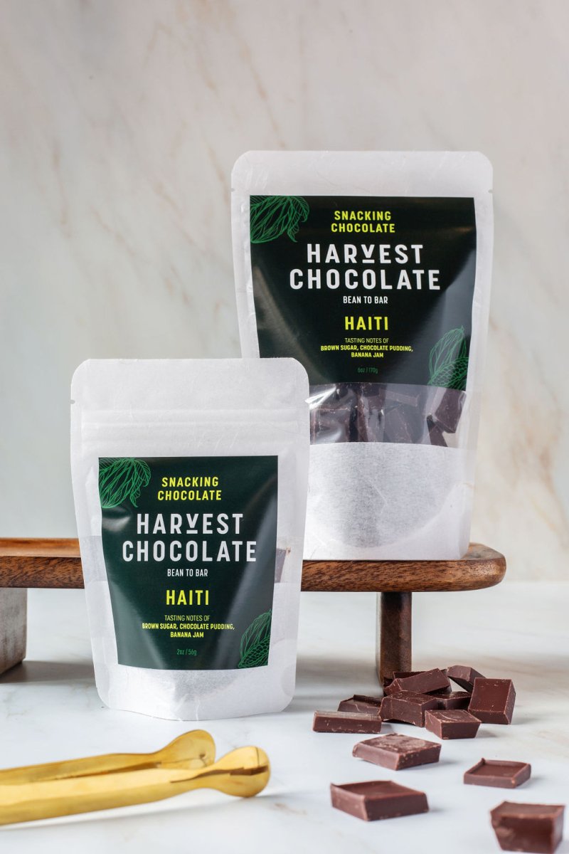 Two pouch sizes of Harvest Chocolate’s Haitian chocolate with snacking pieces on marble surface and gold spoon.