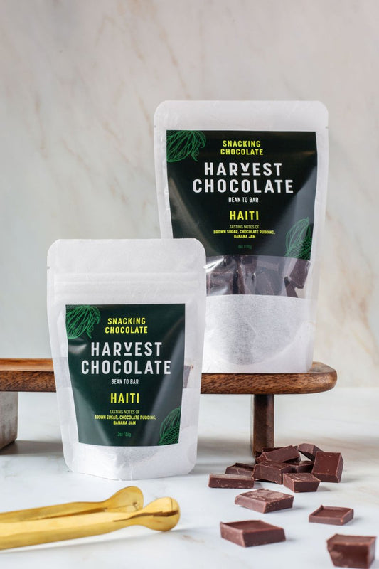 Two pouch sizes of Harvest Chocolate’s Haitian chocolate with snacking pieces on marble surface and gold spoon.