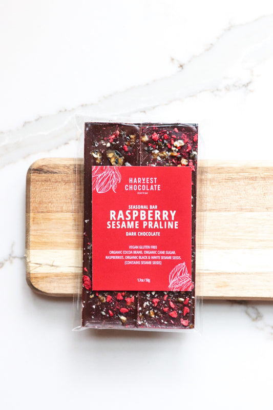 A bar of Harvest Chocolate's Raspberry Sesame Praline dark chocolate, wrapped and displayed on a marble surface with a wooden plank underneath. The packaging features visible raspberry pieces and seeds.