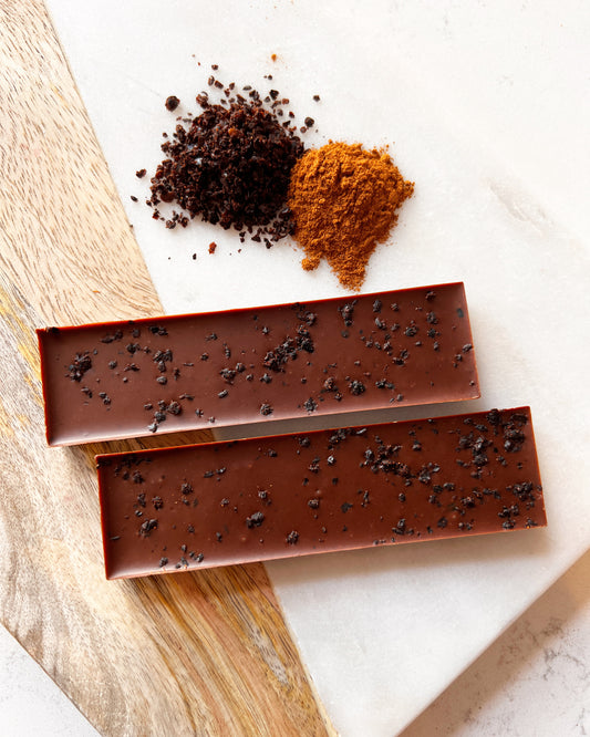 Two bars of dark chocolate sprinkled with coarse salt, positioned on a marble surface beside piles of ground coffee and Harvest Chocolate Urfa Chilli + Cashew. A wooden spoon rests nearby.