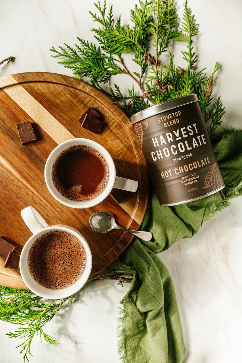 Two mugs of rich hot chocolate made with Harvest Chocolate stovetop hot chocolate mix, styled with greenery on wooden board.