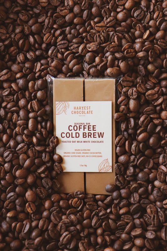 A bar of Harvest Chocolate labeled "Toasted Oat Milk Coffee Cold Brew Chocolate Bar" nestled among a backdrop of scattered coffee beans. The label mentions "seasonal rare," and ingredients such as "to.