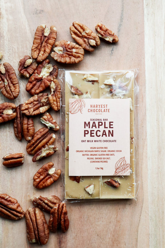 A package of Harvest Chocolate Michigan Maple Pecan chocolate bar with pieces of toasted pecans both inside and scattered around it on a wooden surface. The label includes details of organic and natural ingredients.