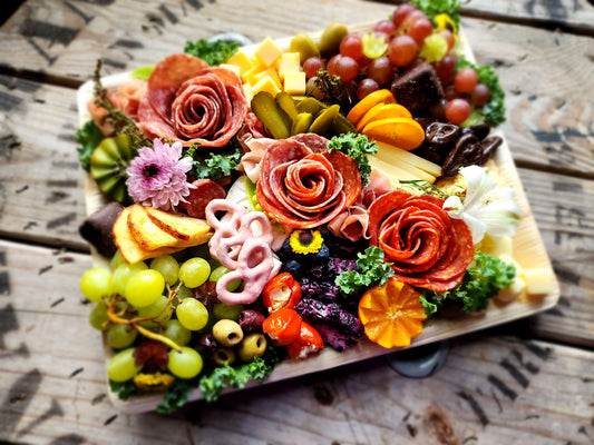 The Charcuterie + Chocolate Workshop by Harvest Chocolate showcases an array of assorted meats, cheeses, olives, grapes, sliced fruits, pickles, and decorative kale. These items are artistically arranged on a wooden surface with flower garnishes and vibrant accents.