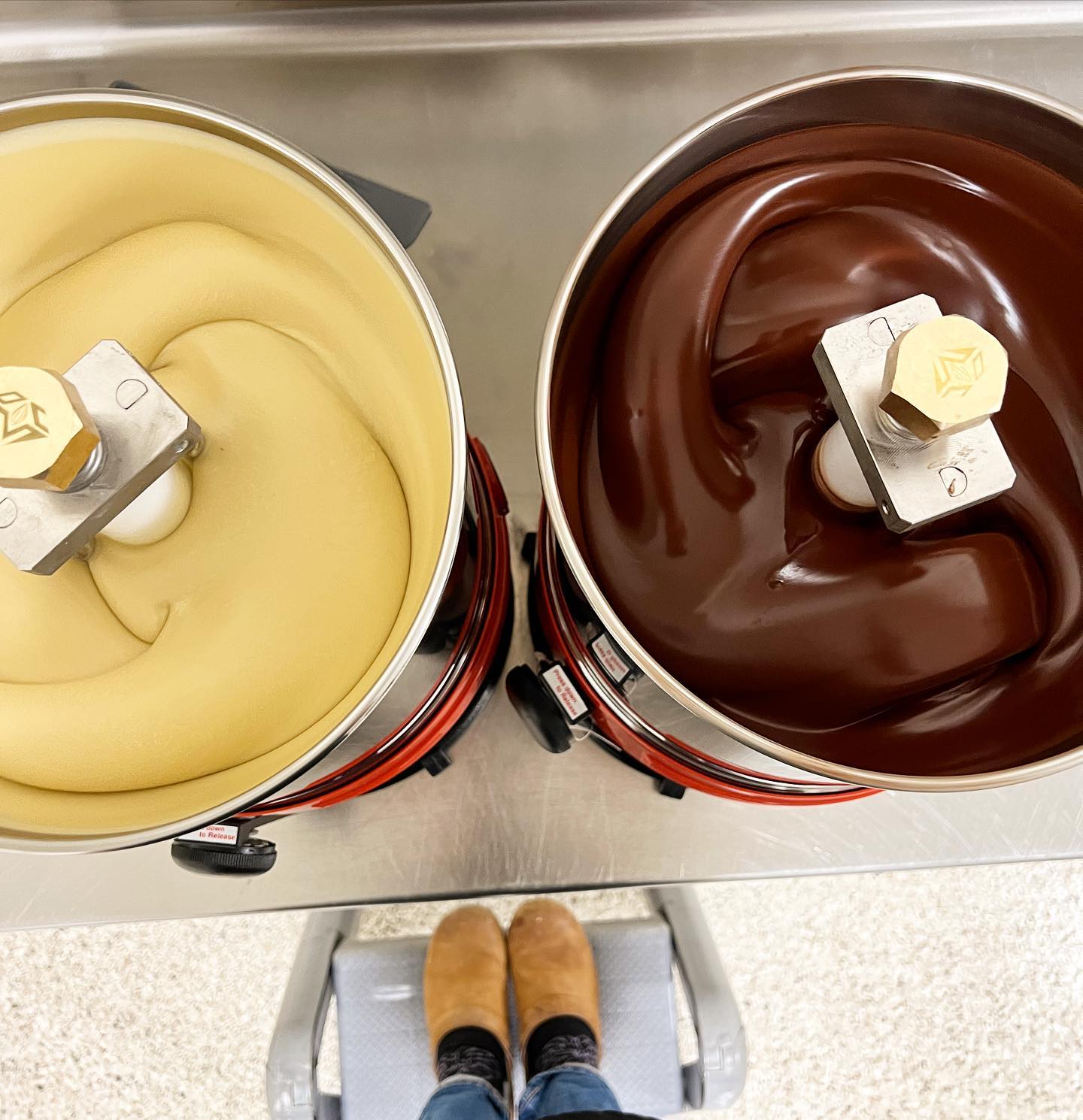 Top-down view of two commercial mixing bowls containing white and milk chocolate, with a person standing between them, seen only from the waist down.