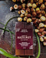 A bar of Harvest Chocolate Hazelnut Milk Chocolate on a surface surrounded by scattered Midwest grown hazelnuts, with a green plant beside it. The label identifies the chocolate as vegan, gluten-free.