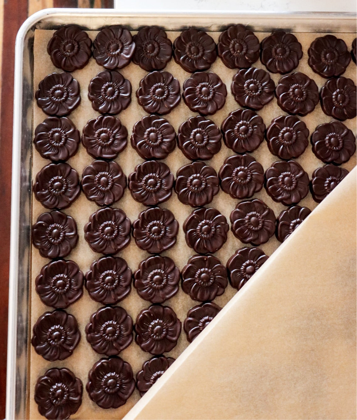 A baking sheet with brown parchment paper displays rows of Harvest Chocolate Flowers by Harvest Chocolate, made from rich Ecuadorian cocoa. Some are partially covered by a folded edge of the parchment.