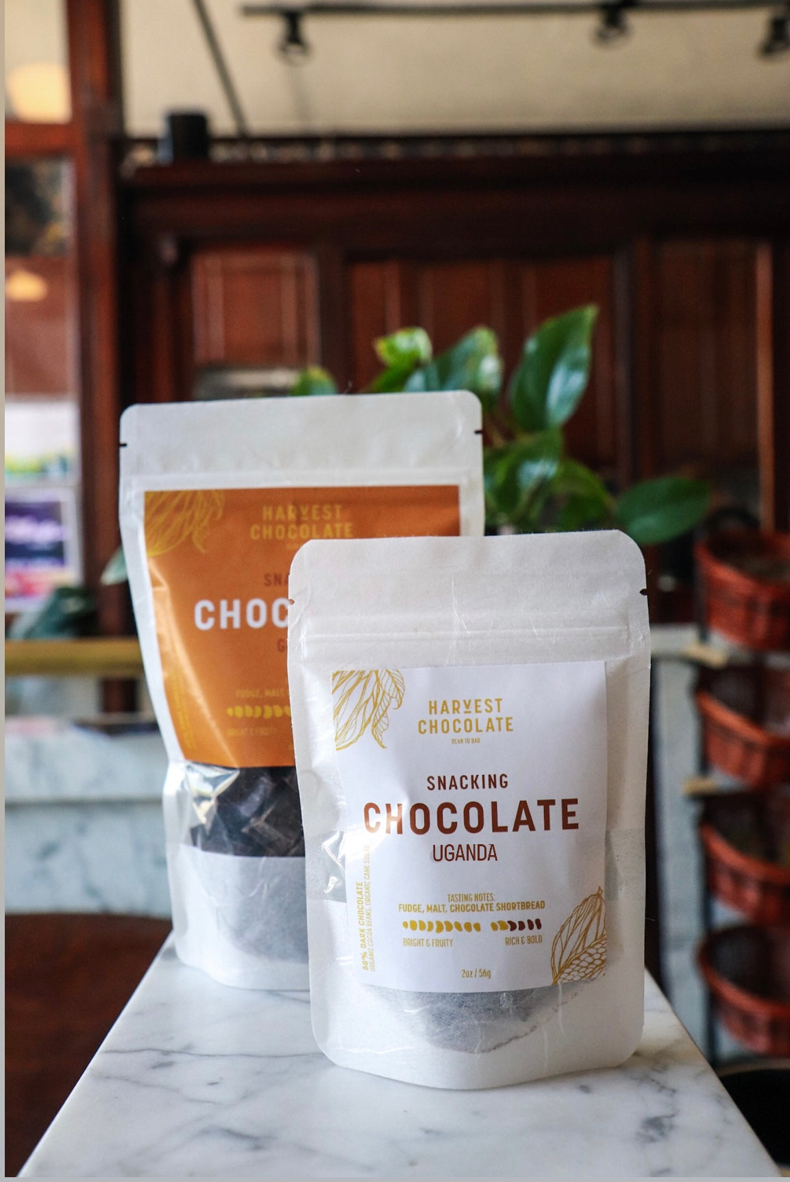 Two bags of Uganda Snacking Chocolate, labeled "Uganda" and "Haiti," standing on a marble counter in a room with wooden accents and green plants in the background.