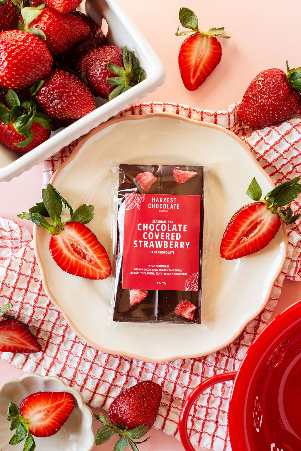 The Harvest Chocolate "Chocolate Covered Strawberry" bar is displayed on a white plate with whole and sliced strawberries. Nearby, a bowl of ripe strawberries and a red dish lie on a pink surface with a checkered cloth, highlighting this vegan treat.