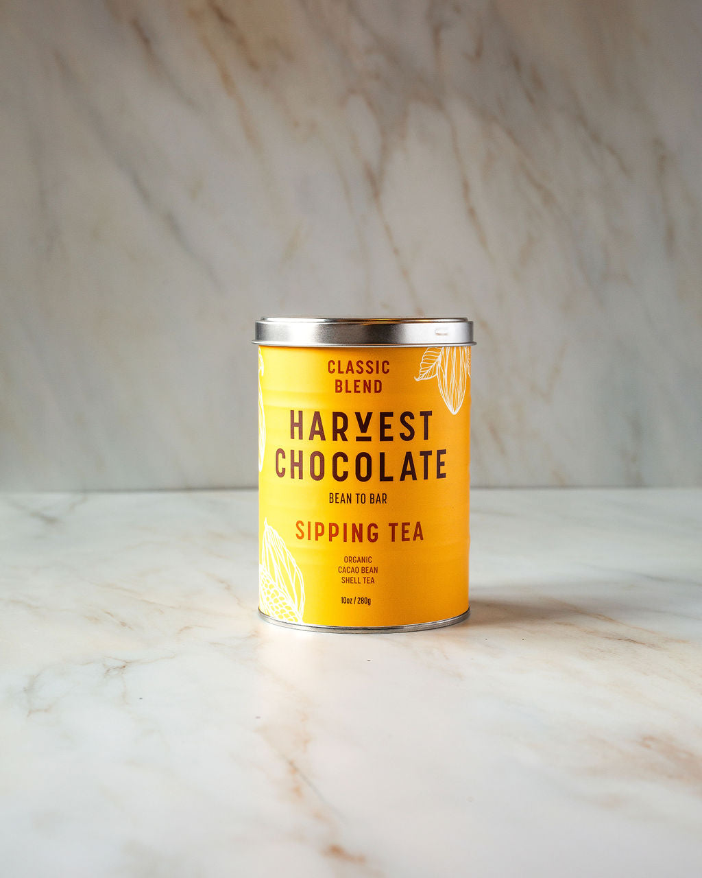 A cylindrical tin with a yellow label rests on a marble surface, featuring the text "Chocolate Tea" and "Organic Cocoa Bean Shell Tea" from the brand Harvest Chocolate. This vegan tea, crafted from organic cocoa bean shells, is topped with a secure metal lid.