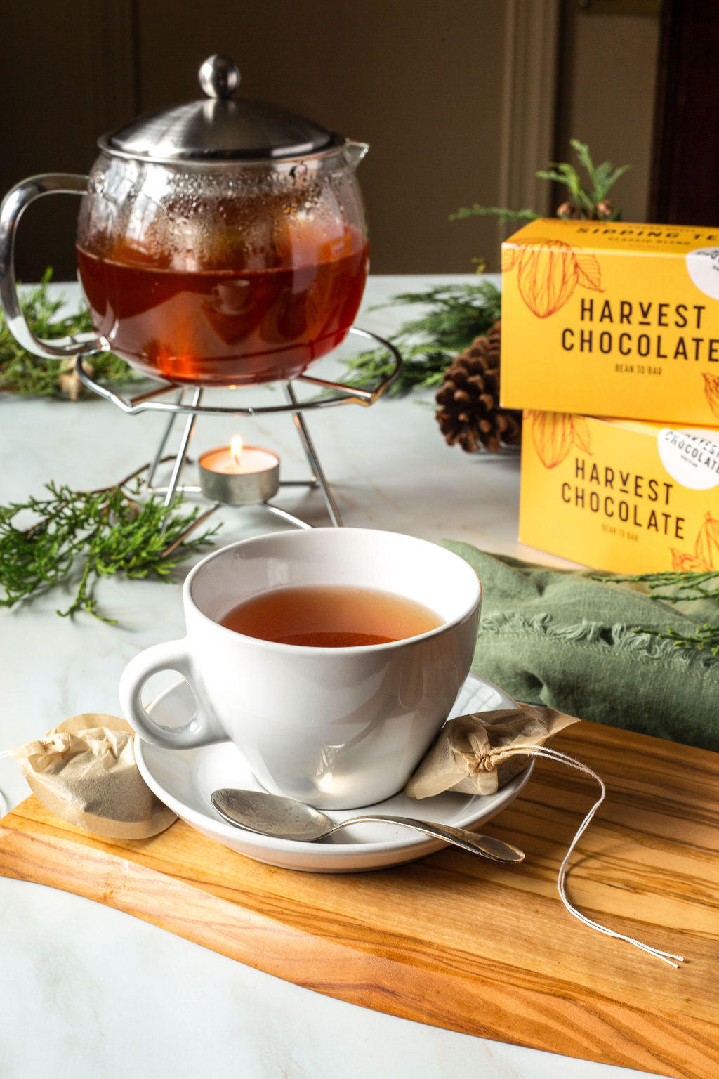 A Harvest Chocolate teacup brimming with Chocolate Tea rests on a saucer, accompanied by a spoon and a teabag. In the background, a glass teapot on a stand holds more looseleaf tea, with the gentle glow of a candle beneath. Boxes of Harvest Chocolate tea and cocoa bean shells enhance the scene on the marble surface.