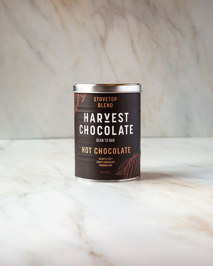 A tin of Harvest Chocolate's Stovetop Hot Chocolate rests on a marble surface. The brown label, featuring white and orange text, is embellished with leaf illustrations that highlight its bean-to-brew craftsmanship.