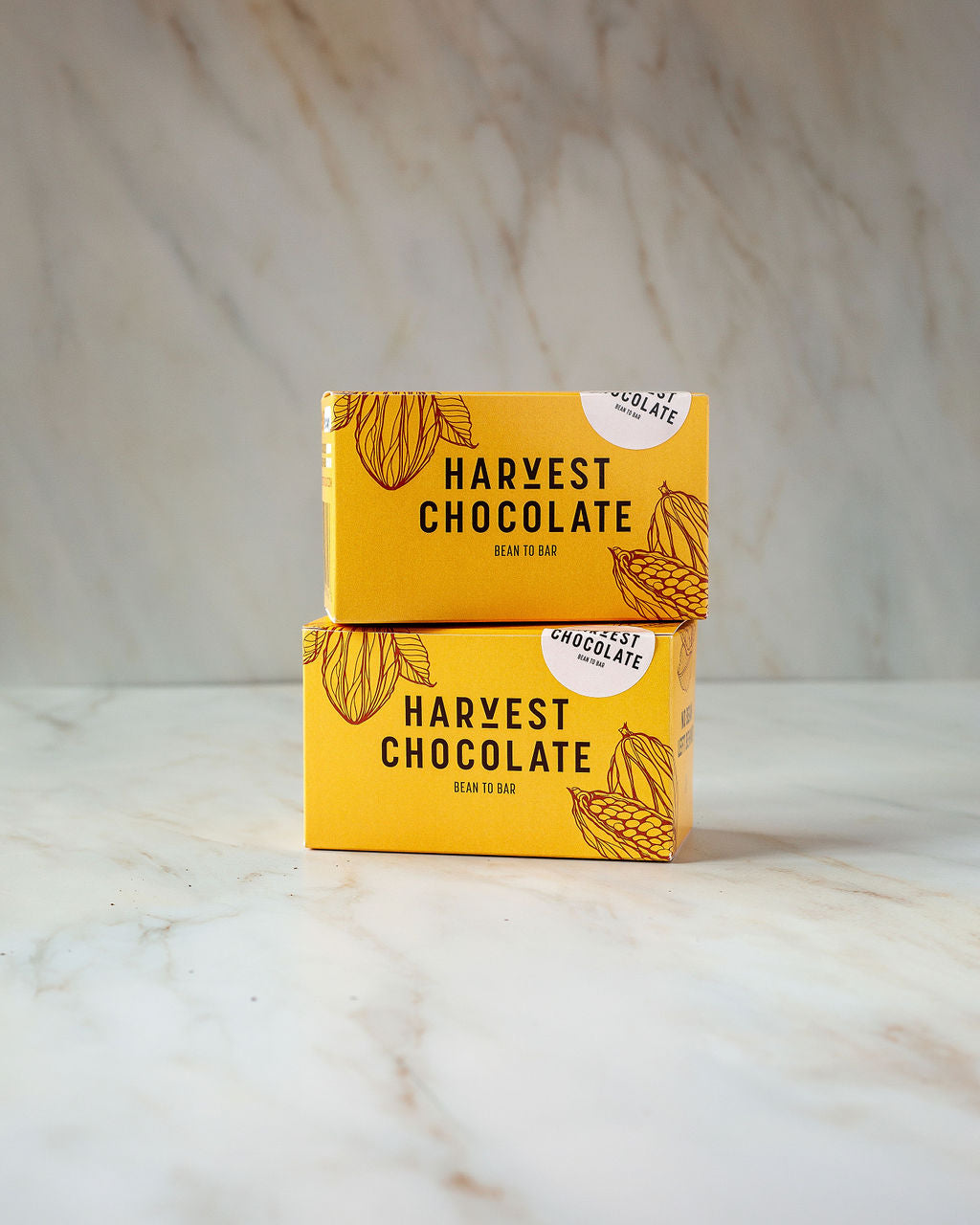 Stacked in an eye-catching arrangement, two yellow boxes labeled "Harvest Chocolate" feature beautiful illustrations of cocoa pods and proudly display the phrase "Bean to Bar." With a commitment to organic ingredients, these Chocolate Tea selections offer a promise of indulgence on a light, marbled surface.