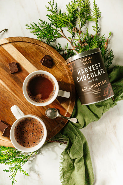 Two cups of Stovetop Hot Chocolate on a wooden tray with small chocolate pieces, accompanied by a Harvest Chocolate canister. Surrounded by green foliage and a green cloth, the scene is complete with a spoon resting beside the cups, promising a rich bean-to-brew experience.