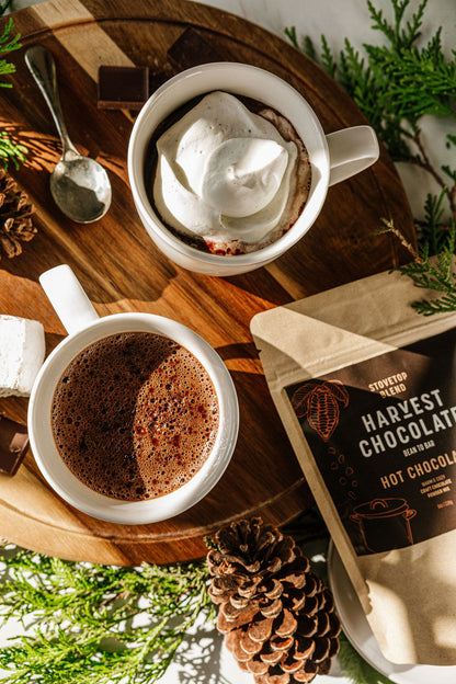 A wooden tray holds two mugs of Stovetop Hot Chocolate from Harvest Chocolate; one mug is topped with whipped cream. Nearby are a spoon, chocolate pieces, and a packet labeled "Bean to Brew Craft Hot Chocolate," emphasizing the inclusion of organic cane sugar. Pine cones and greenery add to the inviting ambiance.