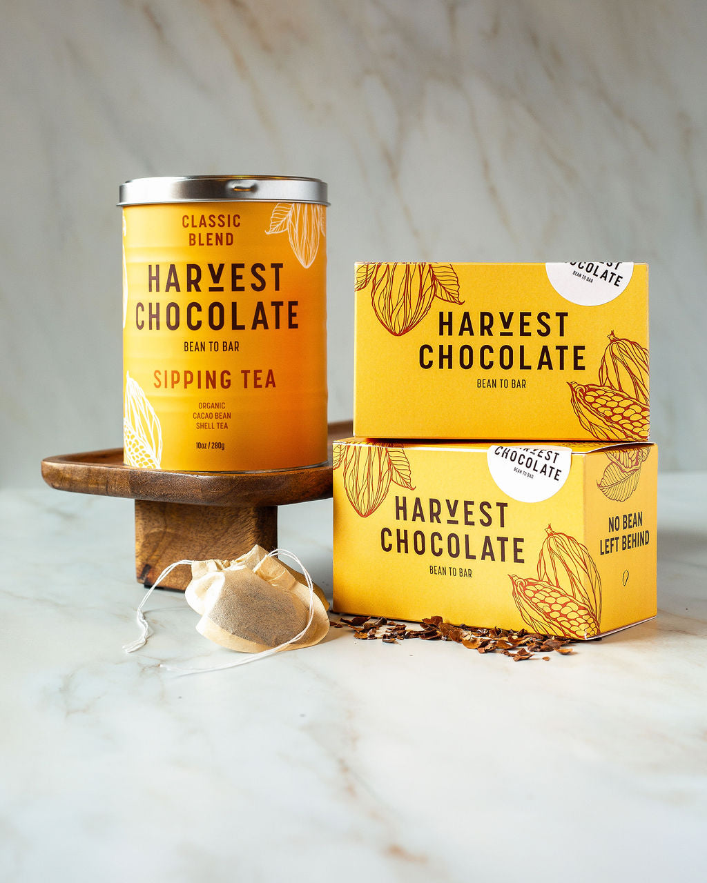 A tin of Chocolate Tea from Harvest Chocolate and two boxes from the same brand rest on a marble surface. The packaging, featuring yellow leaf designs, exudes warmth. A tea bag along with some loose organic cocoa bean shells complete the inviting display.