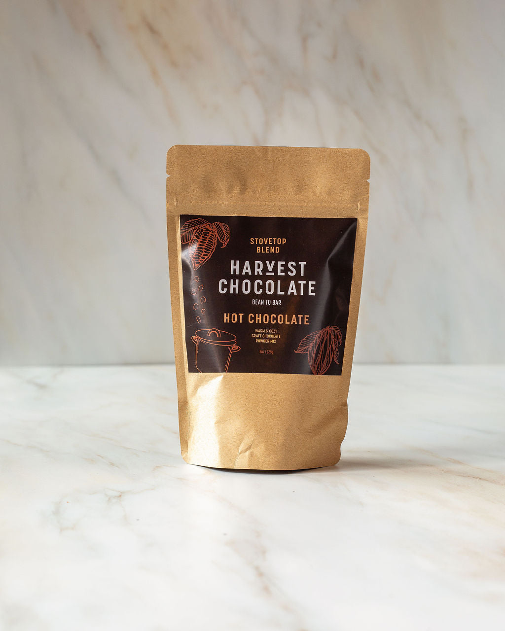 A brown paper bag labeled "Stovetop Hot Chocolate" from Harvest Chocolate sits on a light marble surface. The label boasts "Bean to Brew Craft Hot Chocolate" and "Stovetop Blend," with an illustration of a steaming mug. Made with organic cocoa beans, it's the ideal choice for true chocolate aficionados.