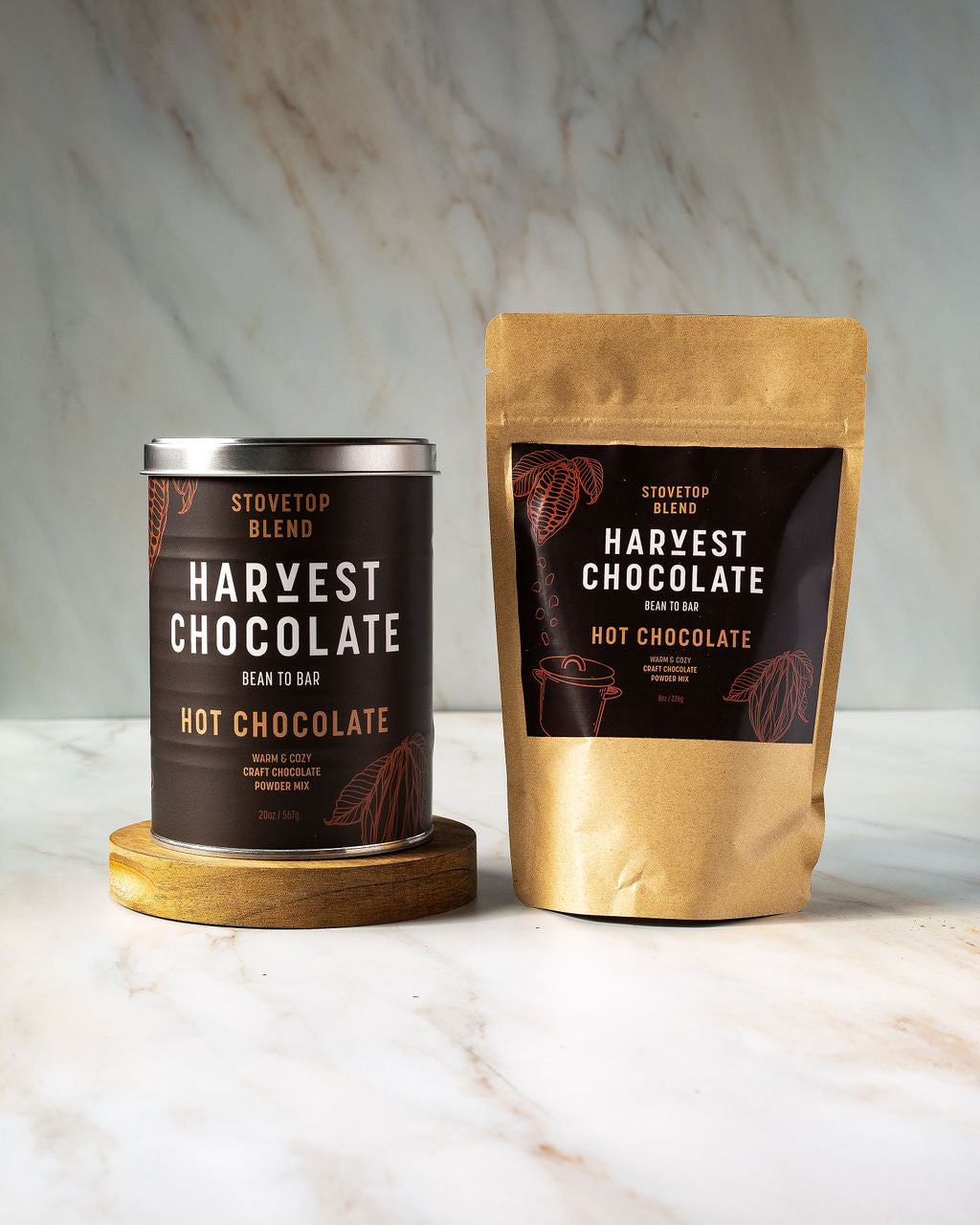 Two packages of "Stovetop Hot Chocolate" from Harvest Chocolate are displayed. One is a cylindrical tin with a lid, and the other is a brown resealable pouch. Both feature dark-themed labels with decorative cocoa illustrations, highlighting the rich, organic hot chocolate that's perfect for any indulgent moment.