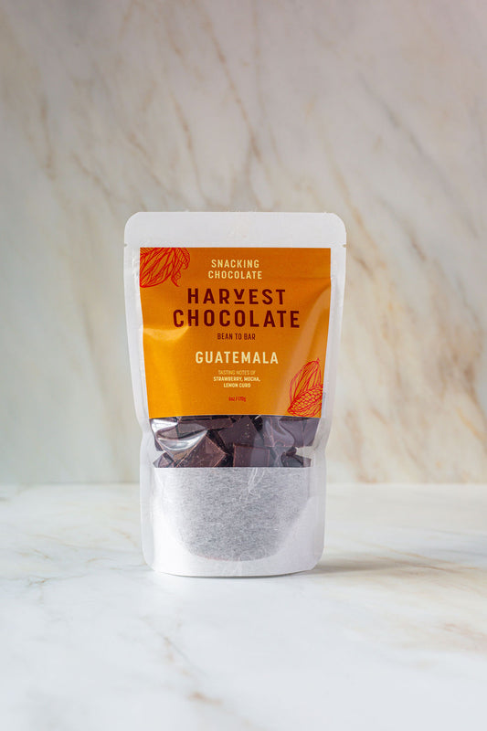 A small white pouch of "Guatemala Snacking Chocolate" from the brand Harvest Chocolate, labeled in orange text, is positioned against a light marble background. The chocolate is noted to be "Bean to Bar," sourced from Lachuá Guatemala.