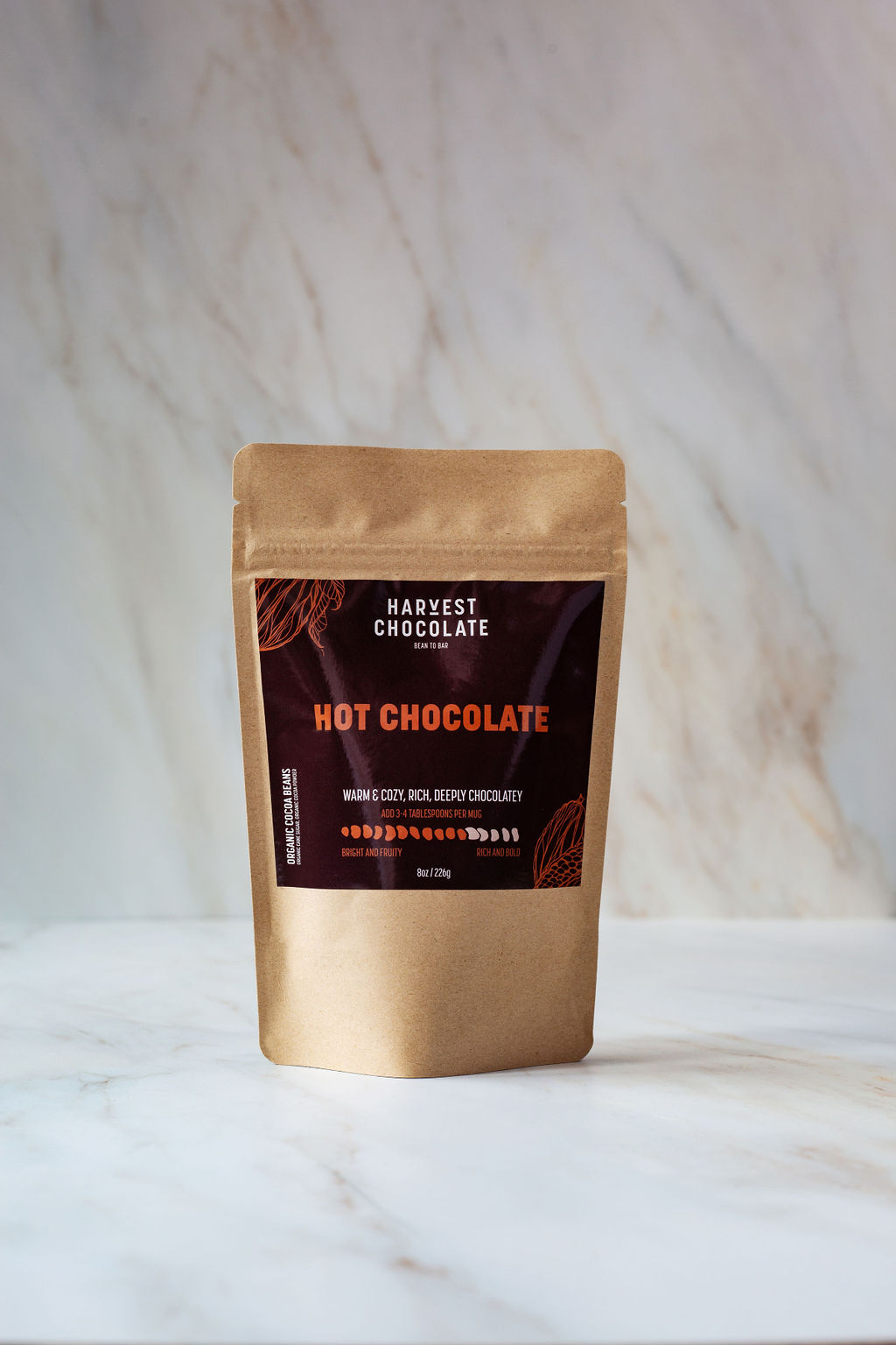 A brown pouch labeled "Hot Chocolate" from Harvest Chocolate is placed on a white marble surface. The simple packaging indicates that it contains dairy-free hot chocolate made with organic cocoa. The surrounding surface is clean and smooth, with subtle brown veining.