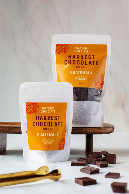 A small white pouch of "Guatemala Snacking Chocolate" from the brand Harvest Chocolate, labeled in orange text, is positioned against a light marble background. The chocolate is noted to be "Bean to Bar," sourced from Lachuá Guatemala.