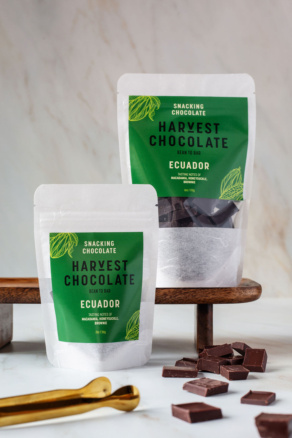 Two bags of "Ecuador Snacking Chocolate" from Harvest Chocolate rest on a wooden board. The front bag, labeled 'bean to bar,' is smaller. Pieces of organic dark chocolate and a gold-colored scoop are arranged on the marble surface in front.