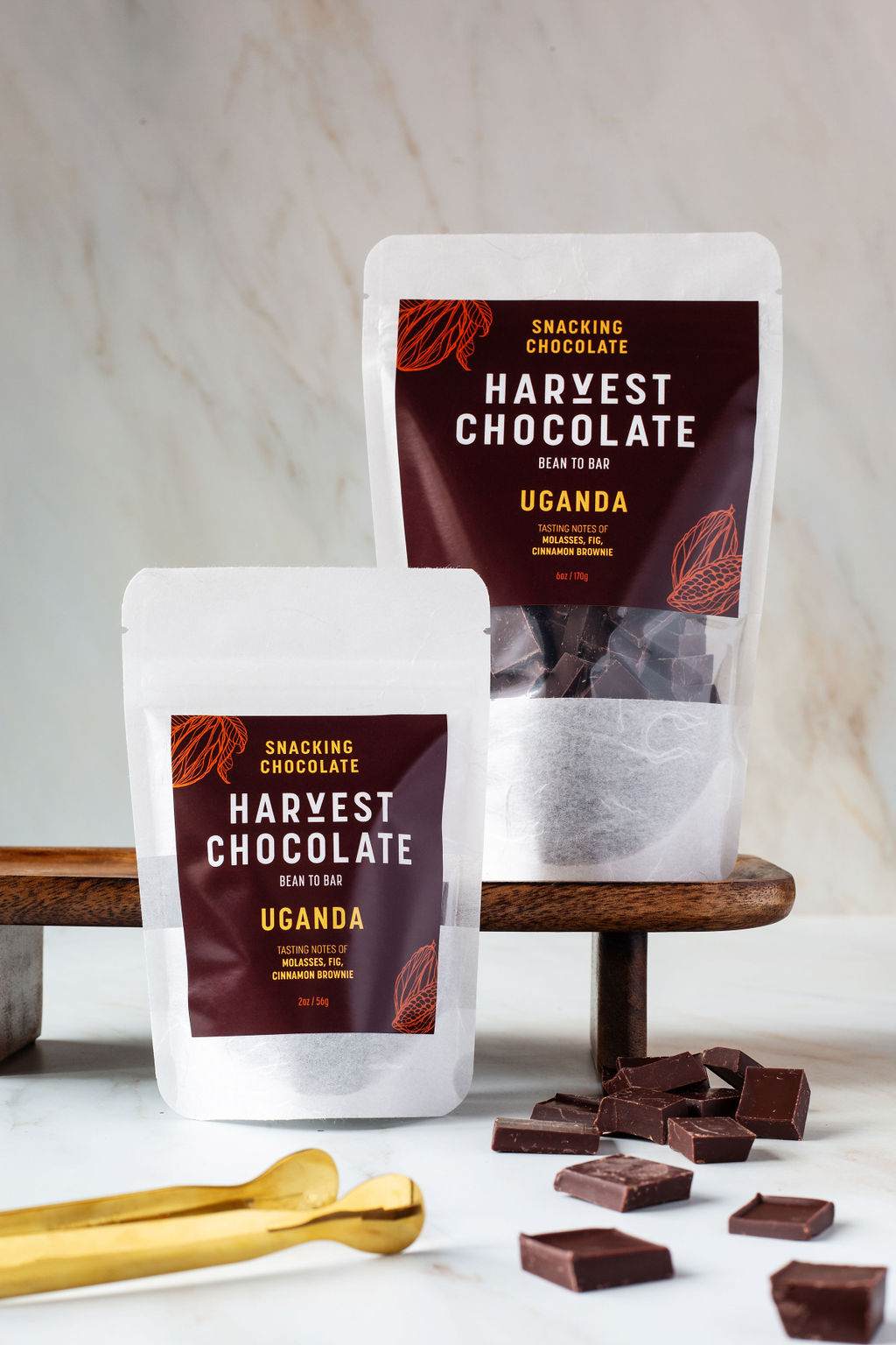 The image displays two white pouches of Harvest Chocolate's Uganda Snacking Chocolate with "Uganda" written on the labels. The pouches are positioned on a dark wooden board, accompanied by scattered pieces of vegan dark chocolate and a gold-colored chocolate breaking tool. The background is a light-colored marble surface, emphasizing their rich fudgy flavor.