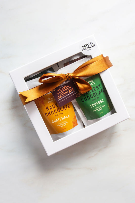 The Travel Through Chocolate Gift Box by Harvest Chocolate contains two bean-to-bar chocolates in a white box, one in yellow packaging labeled "Guatemala" and the other in green packaging labeled "Ecuador." The box is tied with a golden ribbon, embodying a travel-themed chocolate gift.