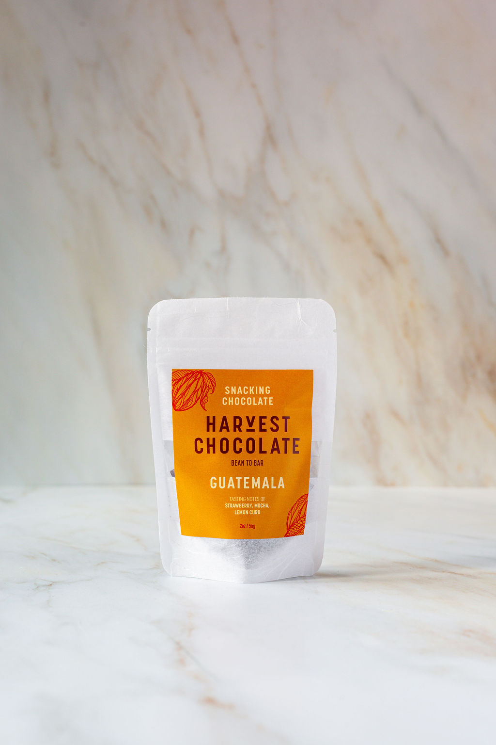 A small white pouch of "Guatemala Snacking Chocolate" from the brand Harvest Chocolate, labeled in orange text, is positioned against a light marble background. The chocolate is noted to be "Bean to Bar," sourced from Lachuá Guatemala.