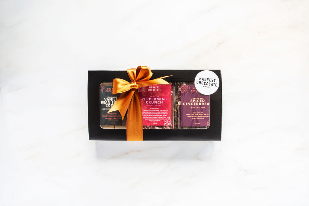 A black box featuring the Holiday Chocolate Bar Trio by Harvest Chocolate includes three distinct bars: Vanilla Bean Sea Salt Coconut, Peppermint Crunch, and Spiced Gingerbread. Tied with a gold ribbon, the box displays a "Harvest Chocolate" sticker, paying tribute to seasonal flavors.