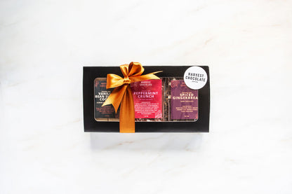 A black box featuring the Holiday Chocolate Bar Trio by Harvest Chocolate includes three distinct bars: Vanilla Bean Sea Salt Coconut, Peppermint Crunch, and Spiced Gingerbread. Tied with a gold ribbon, the box displays a "Harvest Chocolate" sticker, paying tribute to seasonal flavors.