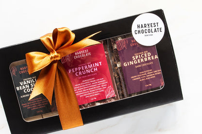 The Holiday Chocolate Bar Trio by Harvest Chocolate, packaged in a black box, includes festive flavors such as Peppermint Crunch, Vanilla Bean Salted Cocoa, and Spiced Gingerbread. The box is adorned with a bronze ribbon and topped with a sticker that reads "Harvest Chocolate Bean to Bar.