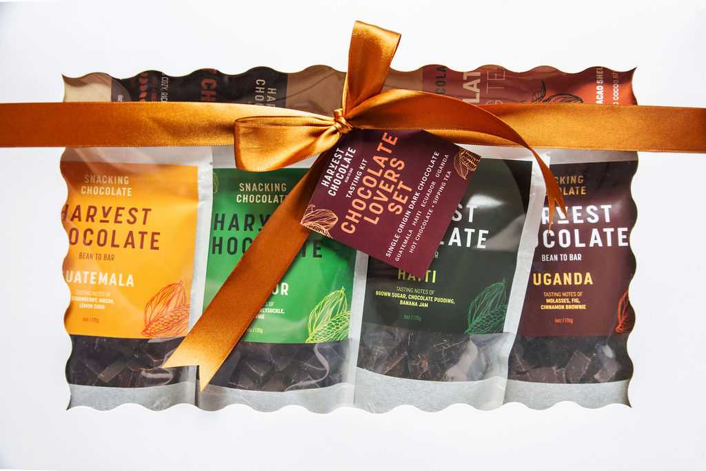 The Chocolate Lovers Gift Box by Harvest Chocolate is beautifully presented in a white box with clear windows, featuring a selection of five delicious snack packs. Each pack showcases different bean-to-bar origins from Guatemala, Honduras, Peru, Haiti, and Uganda. An orange ribbon with a tag elegantly ties around the dark chocolate box.