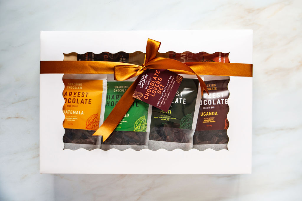 A white Chocolate Lovers Gift Box from Harvest Chocolate, featuring a clear window, displays five bean-to-bar chocolate bars wrapped in vibrant packaging. The box is adorned with an orange ribbon and accompanied by a tag that reads "Chocolate Snacking Set." It elegantly sits on a light marble surface.