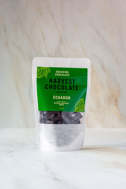 A standing pouch of snacking chocolate labeled "Ecuador Snacking Chocolate" from Harvest Chocolate, displayed against a marble background. The packaging is clear at the bottom, showing chunks of dark vegan chocolate, and green at the top with white and black text detailing the product.