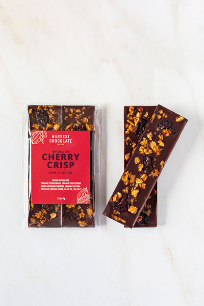 A packaged Cherry Crisp by Harvest Chocolate, featuring Ugandan dark chocolate and dried cherries from Tabone Orchards, is placed on a white, marbled surface. Next to the package are three unwrapped Cherry Crisp bars, topped with visible cherries and crispy bits.