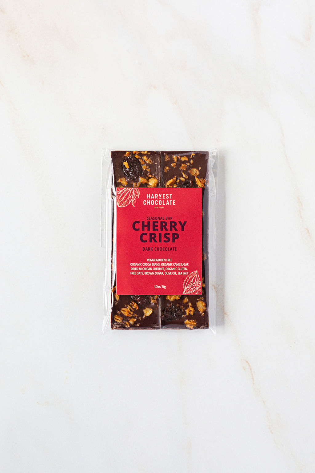 On a white marble surface sits a wrapped chocolate bar labeled "Harvest Chocolate - Cherry Crisp." The clear packaging showcases the dark Ugandan chocolate embedded with dried cherries from Tabone Orchards. The bar weighs 70 grams (2.5 ounces), with the net weight indicated at the bottom of the package.