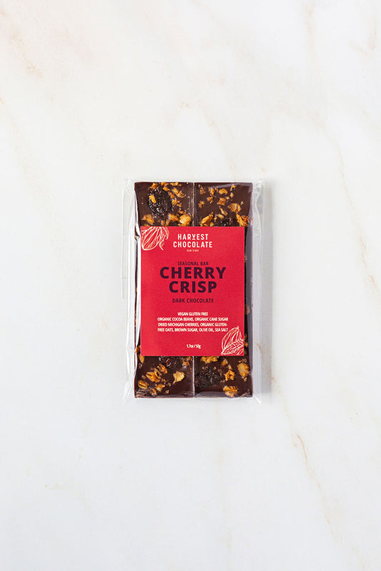 On a white marble surface sits a wrapped chocolate bar labeled "Harvest Chocolate - Cherry Crisp." The clear packaging showcases the dark Ugandan chocolate embedded with dried cherries from Tabone Orchards. The bar weighs 70 grams (2.5 ounces), with the net weight indicated at the bottom of the package.